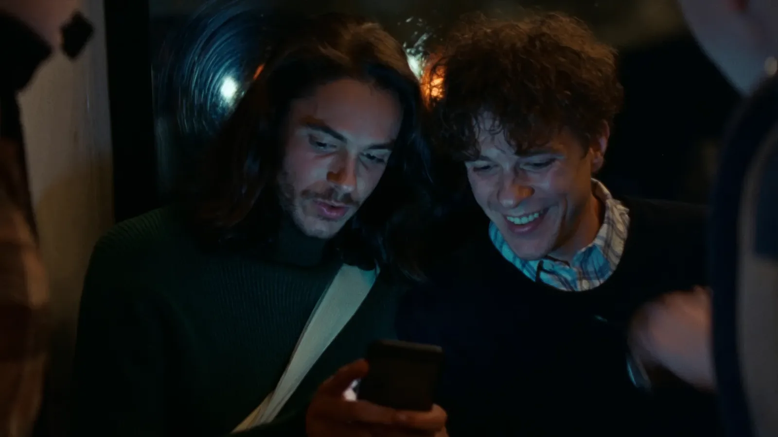 Two friends leaning in over one phone at night, faces lit by the screen, one grinning