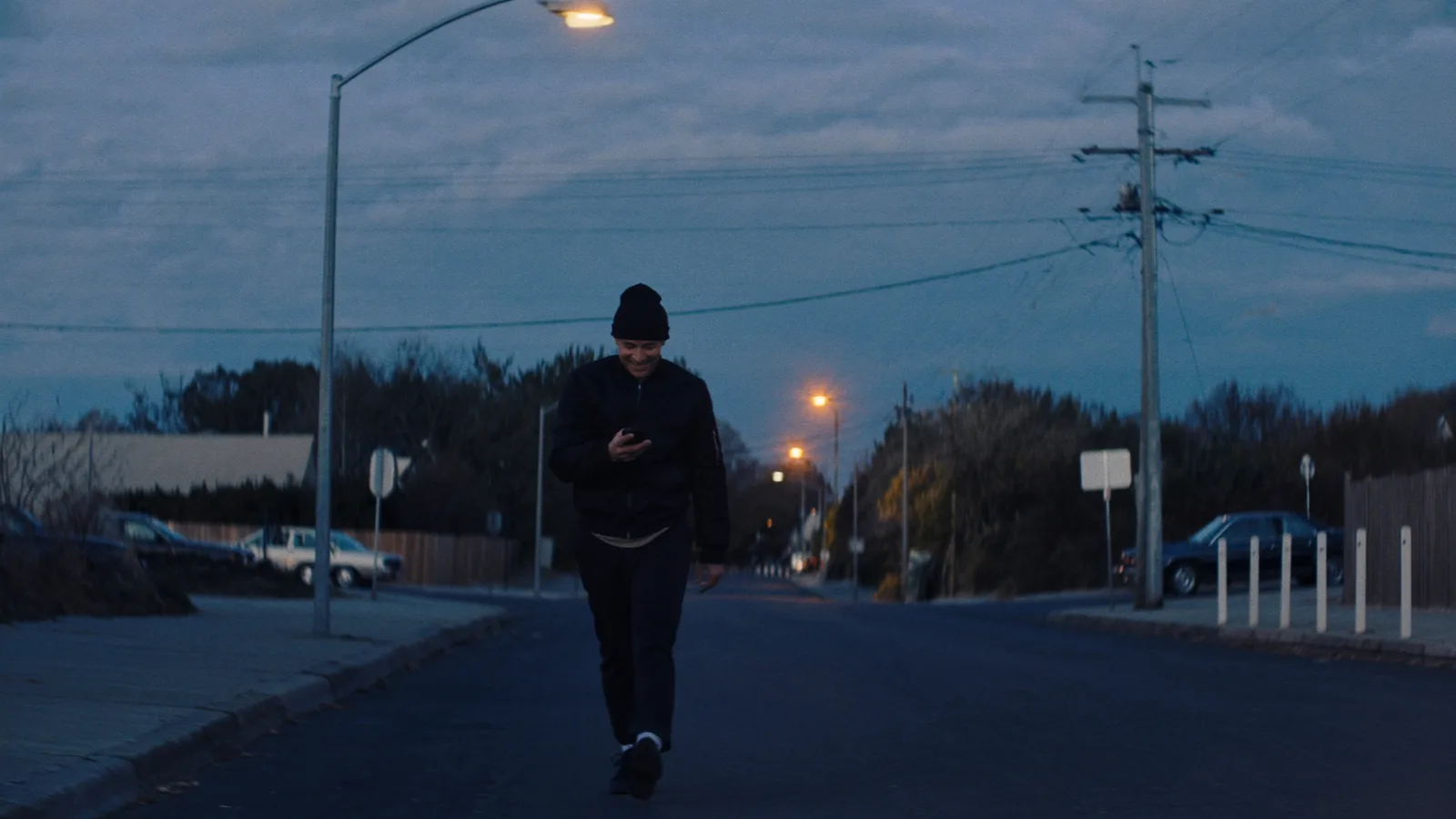 A young man in a beanie walking down an empty street at dusk, smiling at his phone under the streetlights
